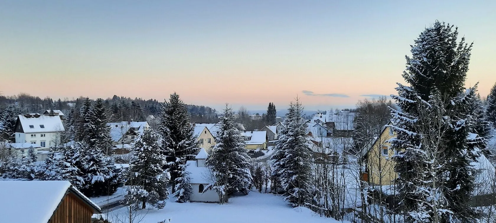Verschneite Dorfdächer und Tannen unter einem zartrosa Abendhimmel, gesehen vom Balkon aus.