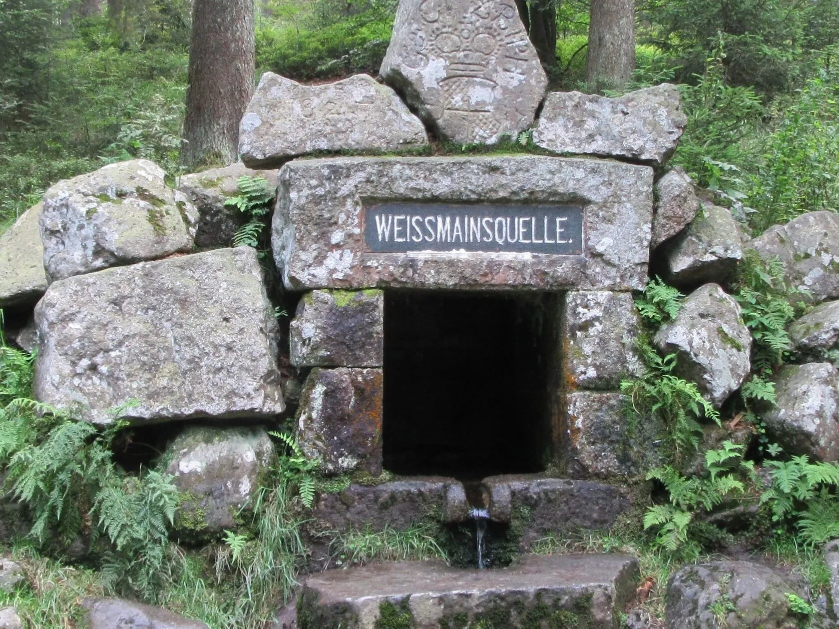 Die steingefasste Weißmainquelle mit der Tafel „Weissmainquelle“, aus der Öffnung rinnt Wasser.