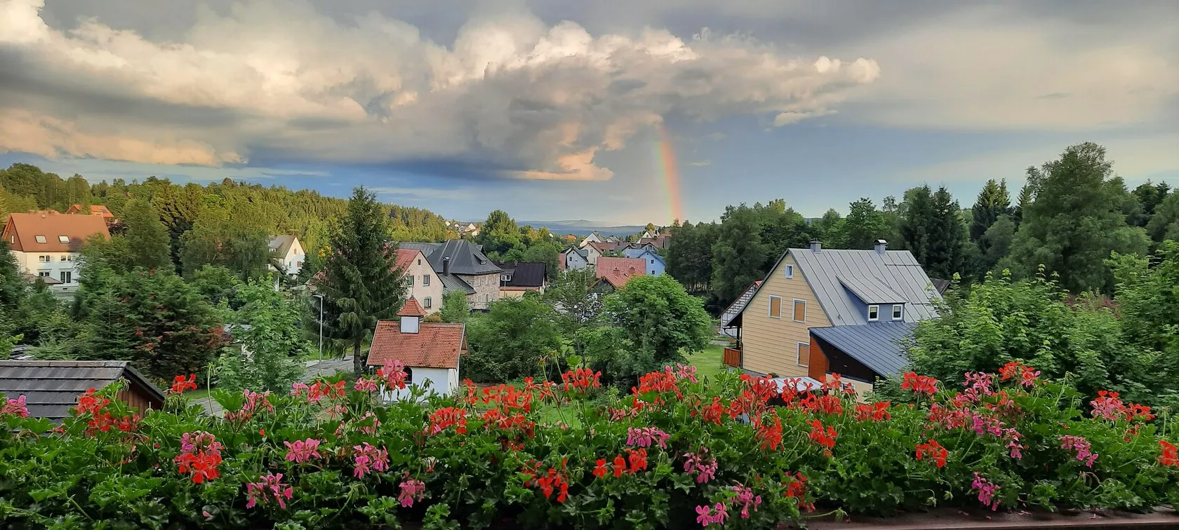 Blick vom Balkon über das Dorf: ein Regenbogen unter aufziehenden Wolken, im Vordergrund rote Geranien.
