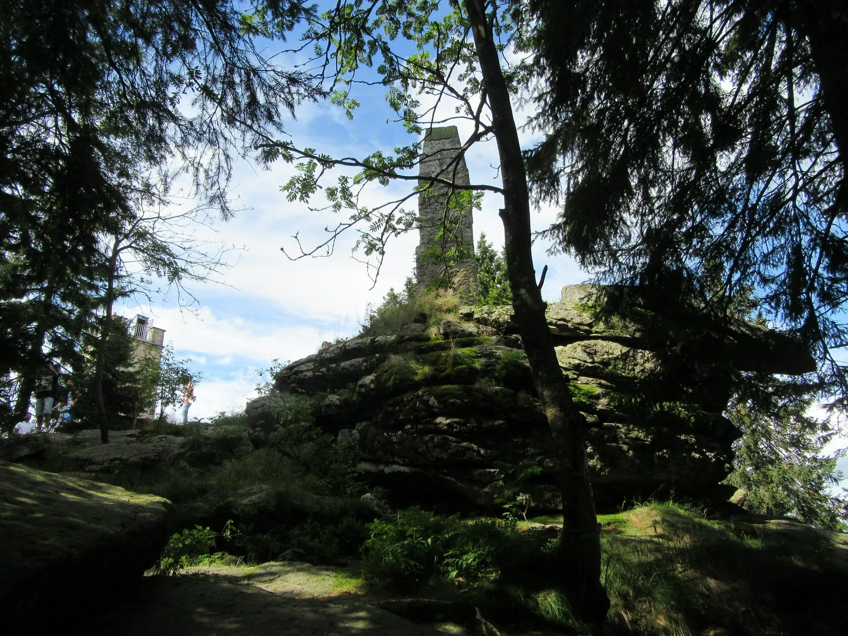 Ein steinerner Turm auf einem Felsgipfel, gerahmt von den Zweigen dunkler Fichten.