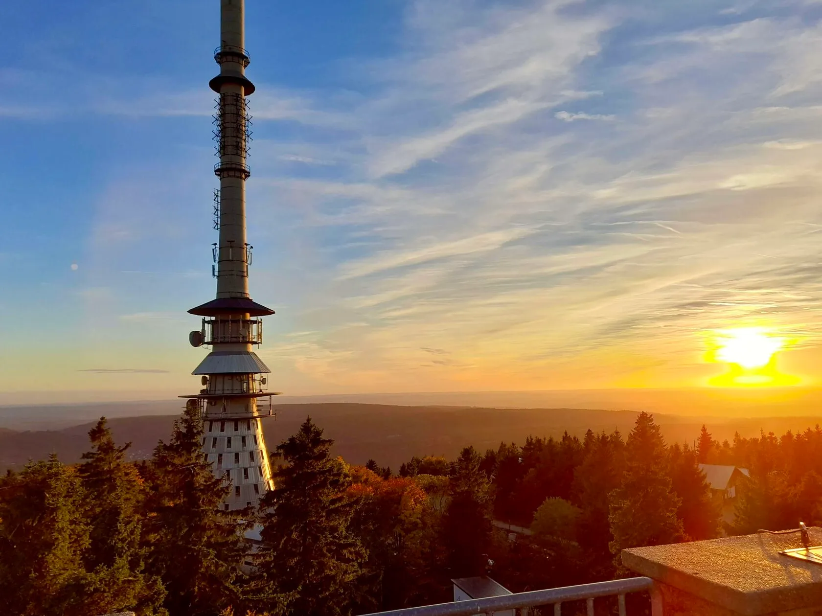 Ein hoher Sendeturm über herbstlich gefärbtem Wald, dahinter die untergehende Sonne über bewaldeten Hügeln.