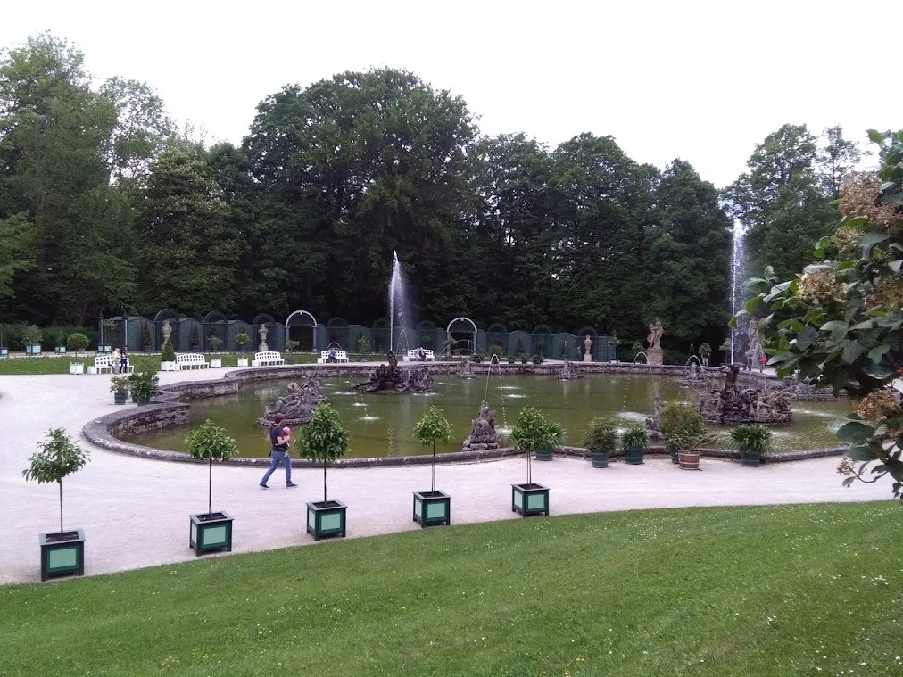 Barockes Wasserbecken mit Fontänen, Steinfiguren und Kübelpflanzen in einem Schlosspark.