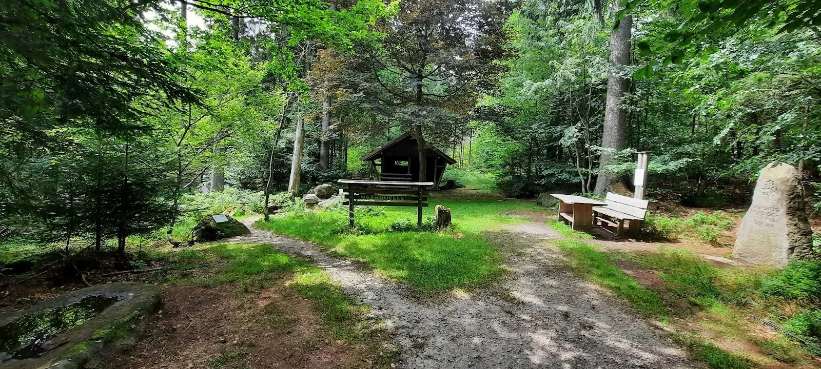 Hölzerne Schutzhütte mit Bänken auf einer Waldlichtung, rechts am Weg ein großer Findling.
